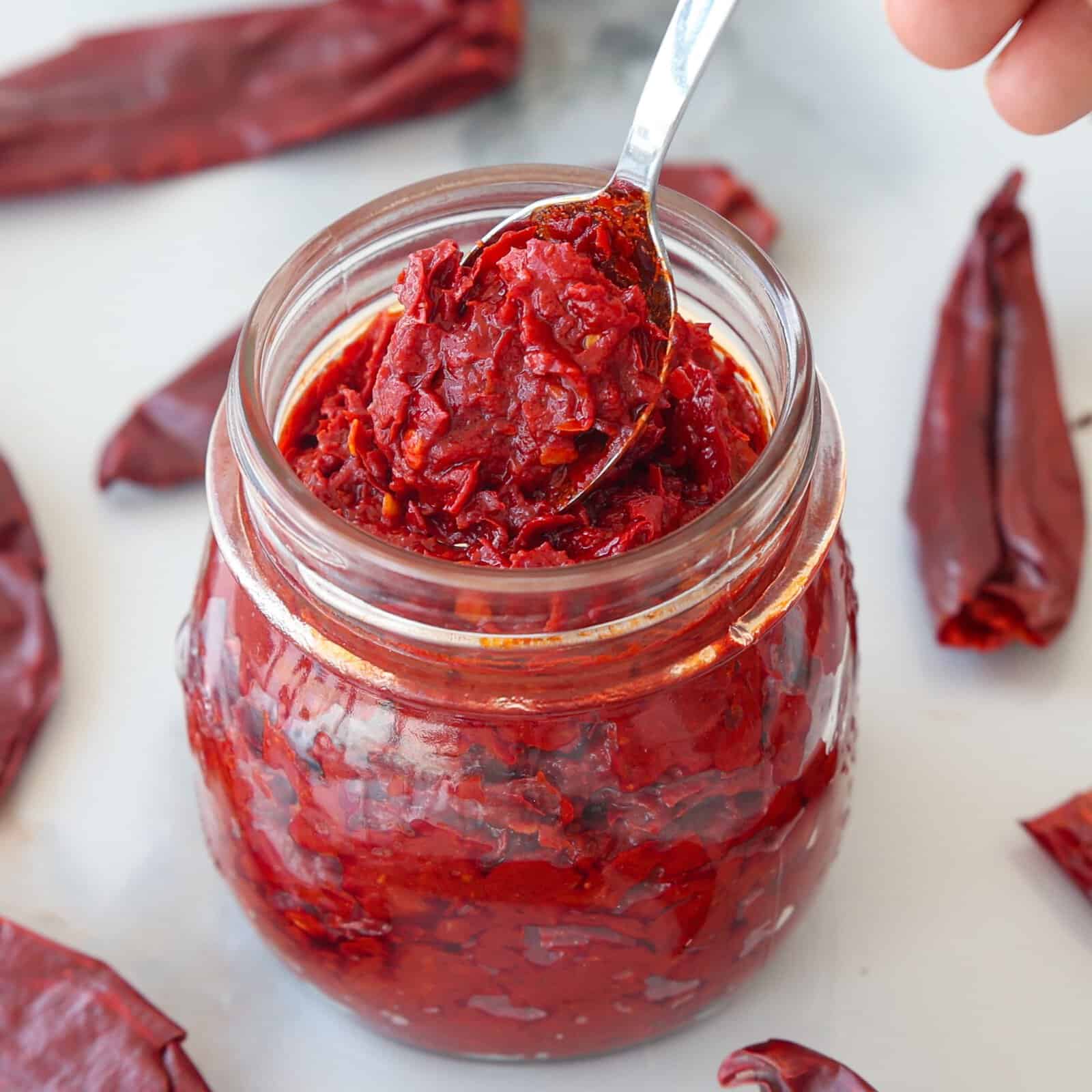 A spoon scoops out thick, vibrant harissa paste from a glass jar, with dried red chilies scattered around on a white surface.