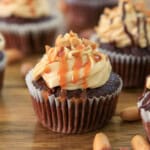 A snickers cupcake topped with creamy frosting, chopped peanuts, and caramel drizzle, sitting on a wooden surface with more cupcakes and peanuts around it.