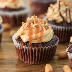 A snickers cupcake topped with creamy frosting, chopped peanuts, and caramel drizzle, sitting on a wooden surface with more cupcakes and peanuts around it.