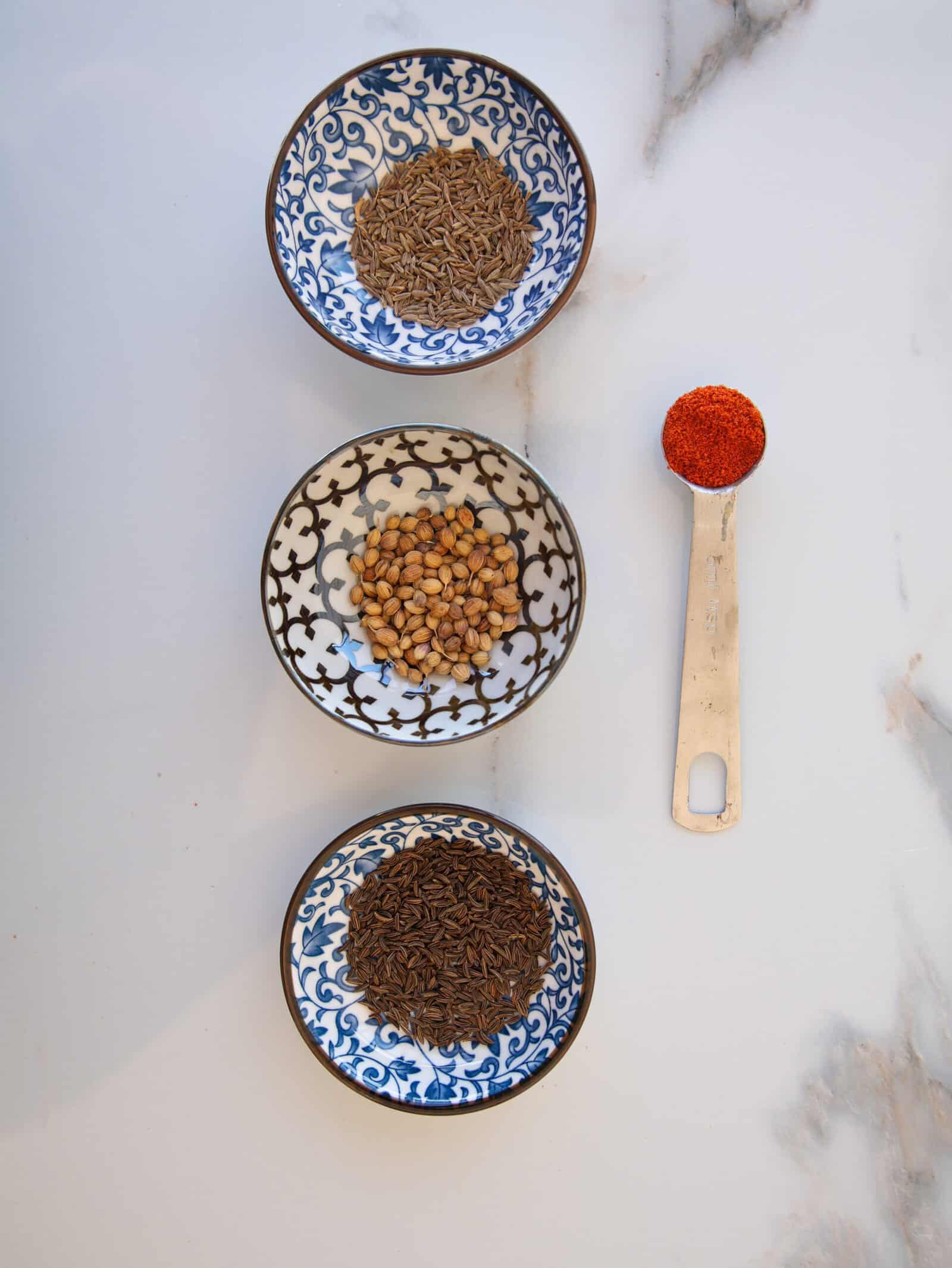 Three ornate bowls on a white surface, containing cumin seeds (top), coriander seeds (middle), and caraway (bottom), with a metal spoon holding paprika to the right of the bowls.