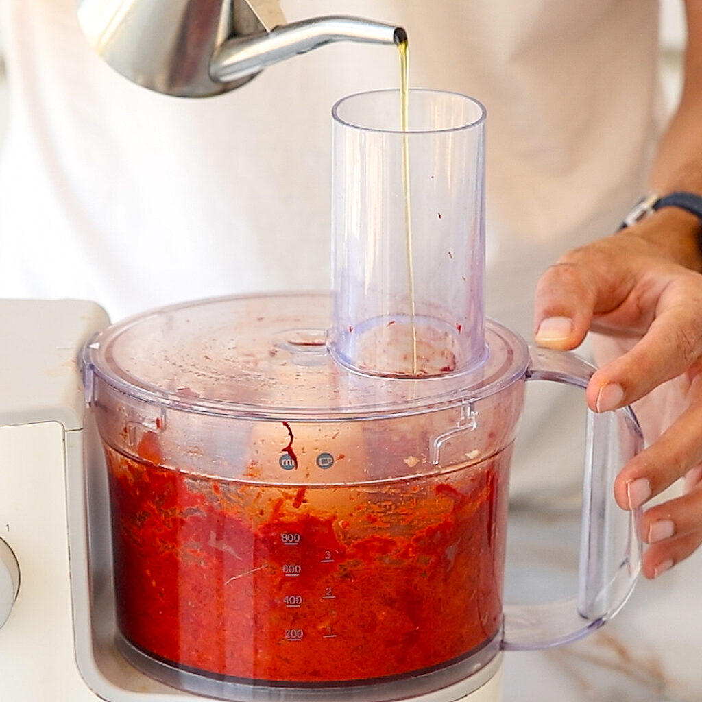A person pours oil into a food processor blending a red sauce or mixture. The food processor is partially filled and the person's hand holds the handle.