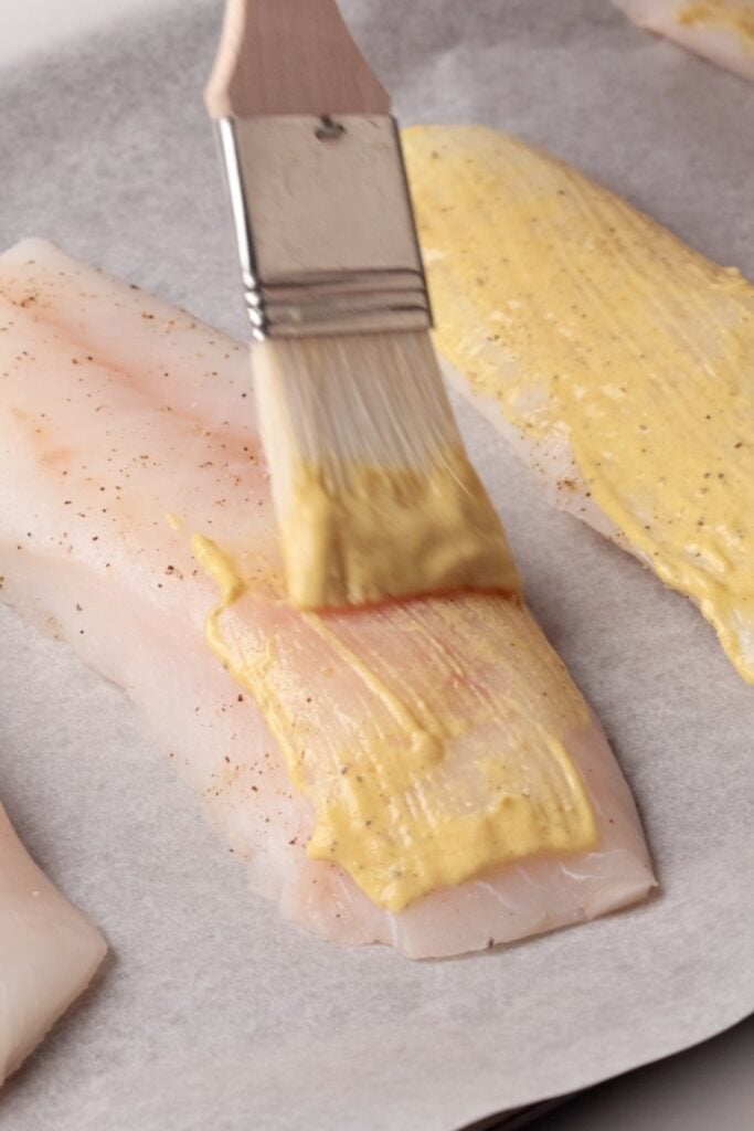 A close-up of a raw codfillet on parchment paper being brushed with a mustard-lemon mix using a small basting brush. Another fillet, already coated with sauce, is nearby.