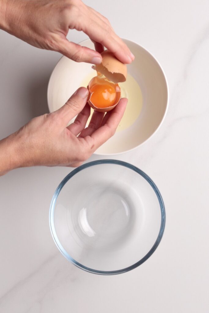 A person cracks an egg, separating the yolk and egg white over a white bowl, with an empty glass bowl below on a white countertop.