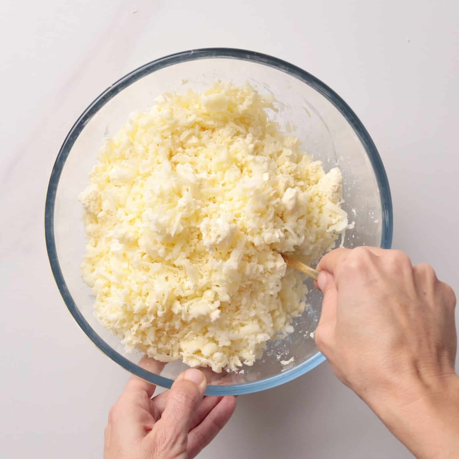 A person mixing grated cheese for khachapuri in a clear glass bowl using a wooden spoon on a white surface.