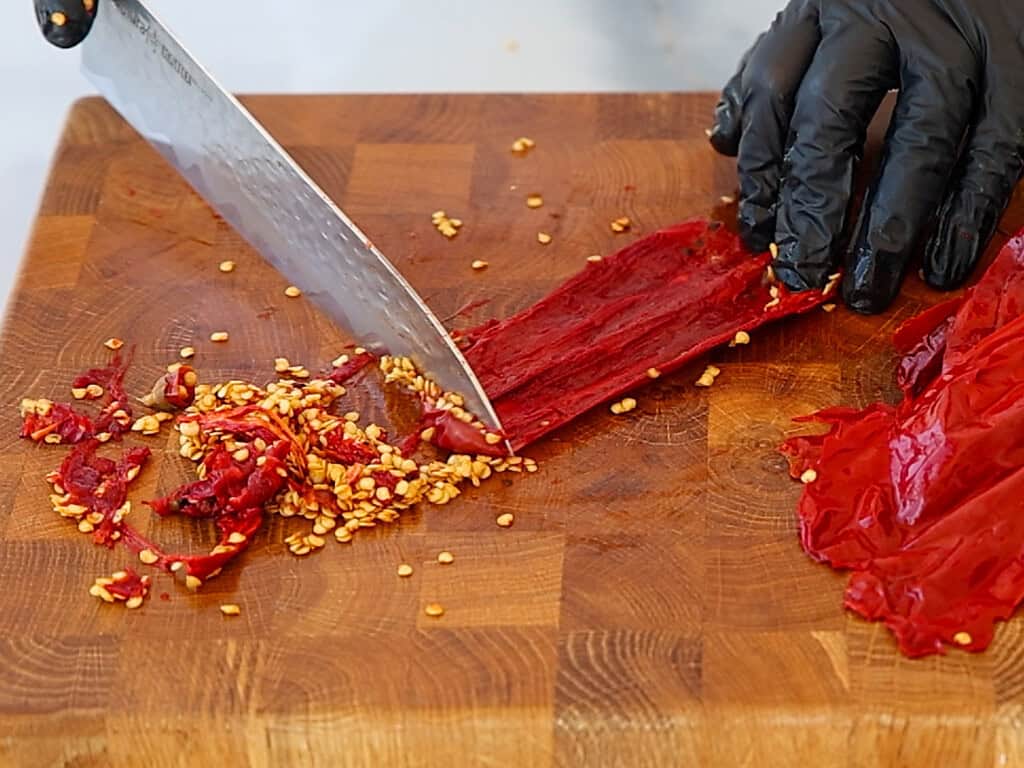 A person wearing black gloves slices a red chili pepper on a wooden cutting board, with seeds and pepper pieces scattered around.