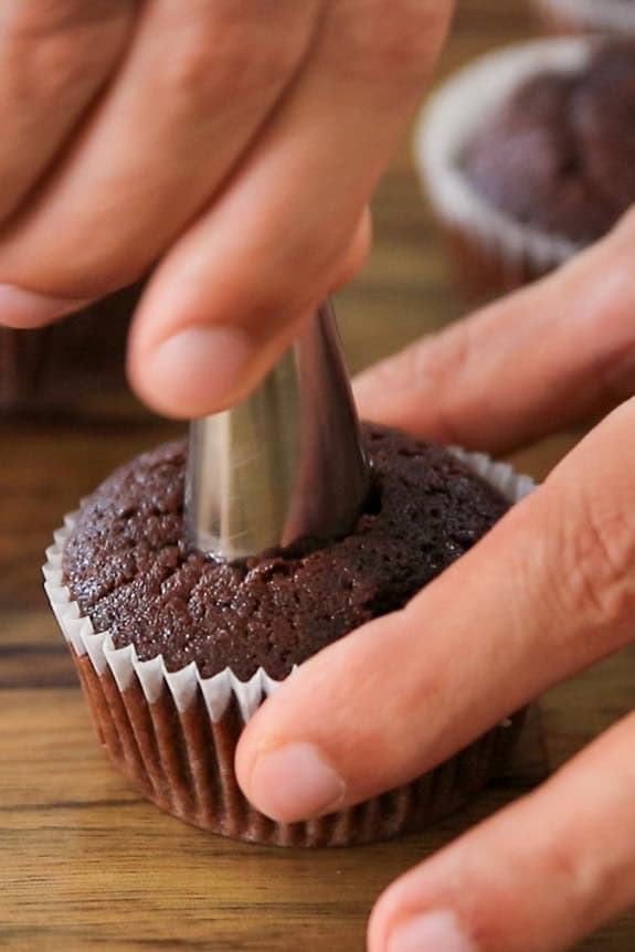 Close-up of hands using a metal piping tip to core a chocolate cupcake in a white paper liner on a wooden surface.