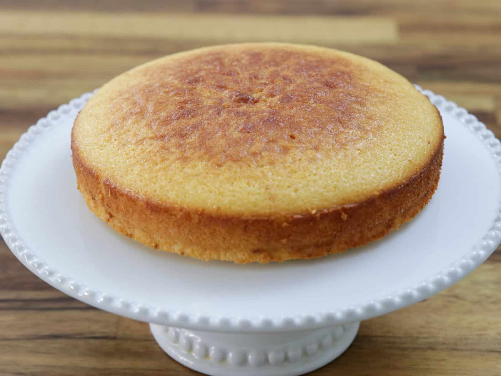 A round, golden-brown 4-ingredient condensed milk cake sits on a white, decorative cake stand with a wooden surface in the background.