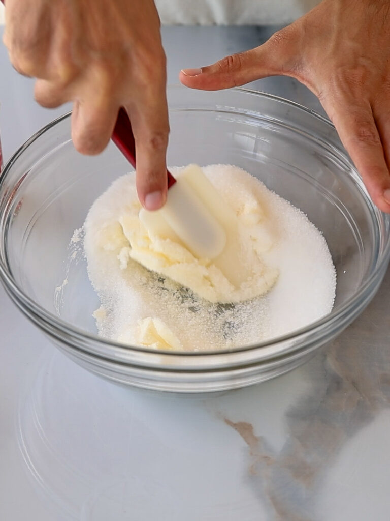 A person uses a spatula to mix butter and sugar in a clear glass bowl on a light surface.
