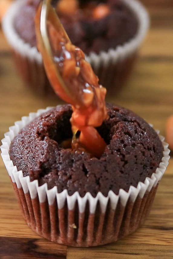 A chocolate cupcake with a hollowed center in a paper liner is being filled with gooey caramel sauce from a spoon. Another cupcake is blurred in the background on a wooden surface.