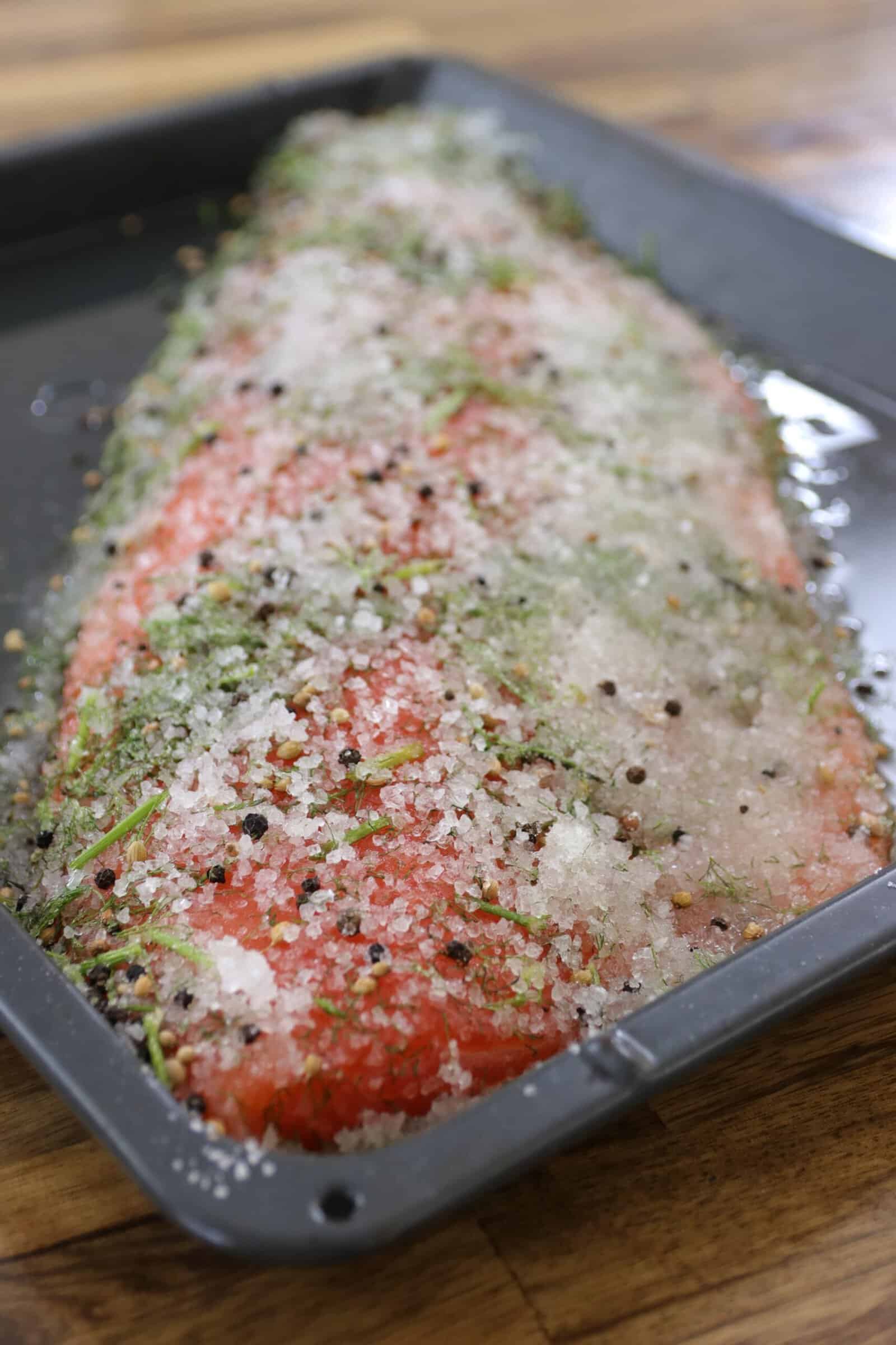 A raw salmon fillet in a baking tray, coated with a mixture of coarse salt, herbs, and peppercorns, preparing to be cured or baked, on a wooden surface.