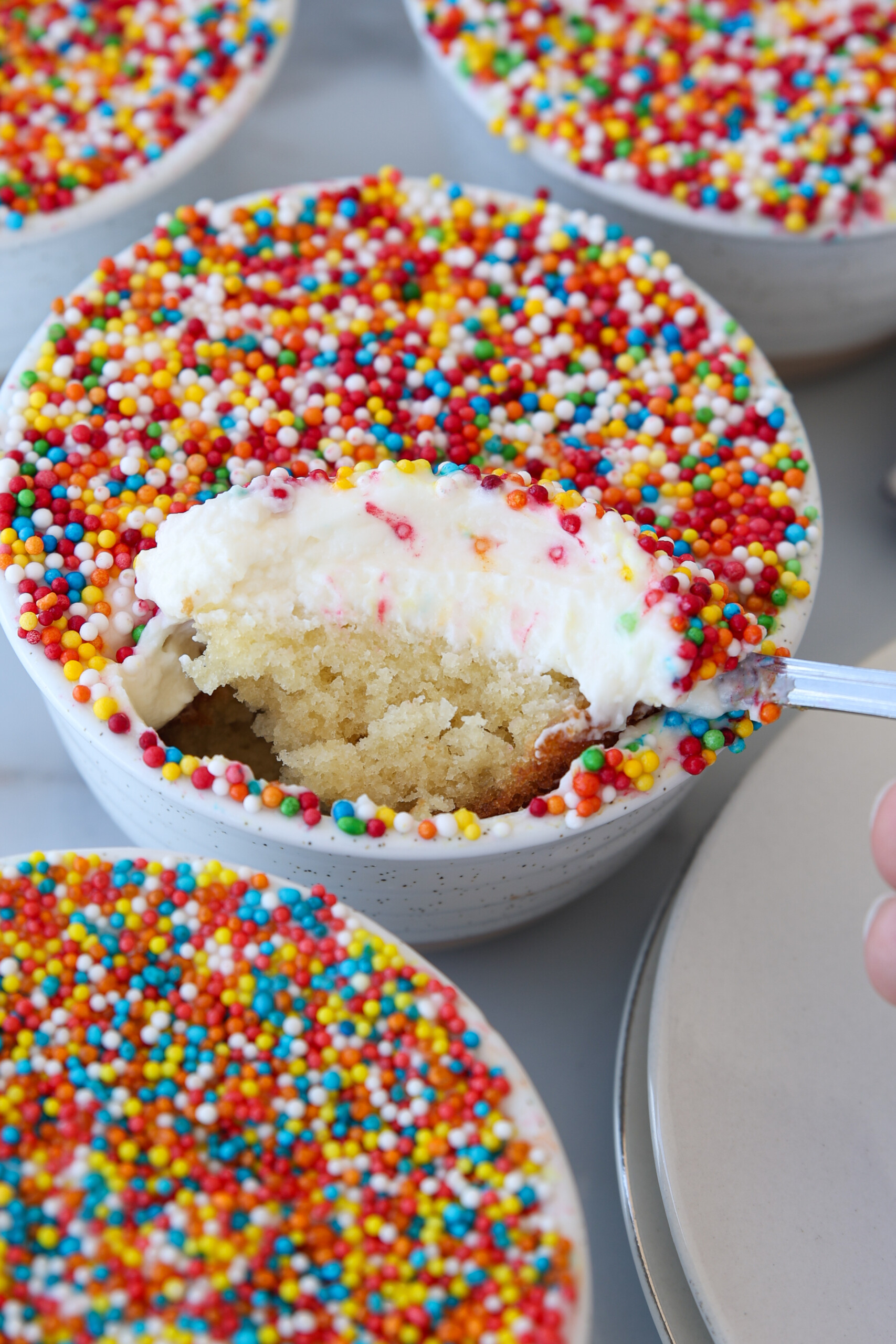 A spoon scoops vanilla dot cake with white frosting and colorful round sprinkles from a small white bowl, surrounded by other identical sprinkle-covered cakes.