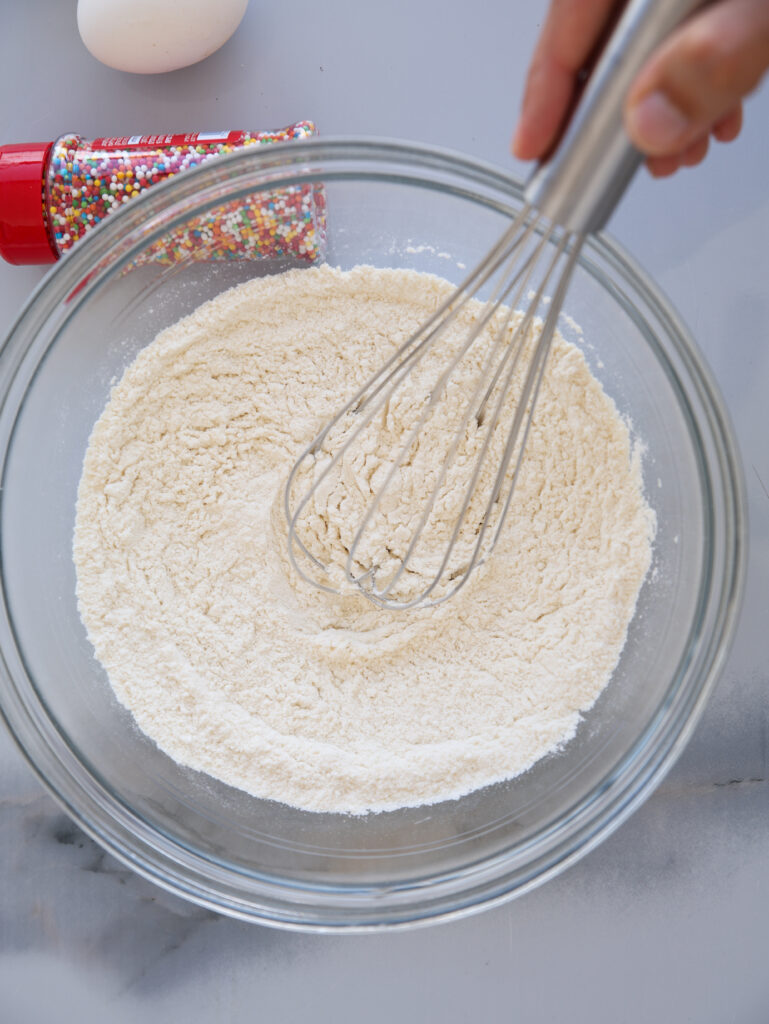 A hand holds a whisk stirring flour in a glass bowl. Nearby, a bottle of colorful sprinkles and an egg rest on the gray countertop.