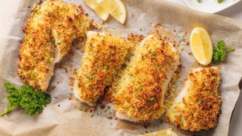 Baked Panko Crusted Cod Four pieces of baked, panko crusted cod fish fillets on parchment paper, garnished with fresh parsley and lemon wedges. The fish has a golden, crispy topping and is presented on a baking tray.