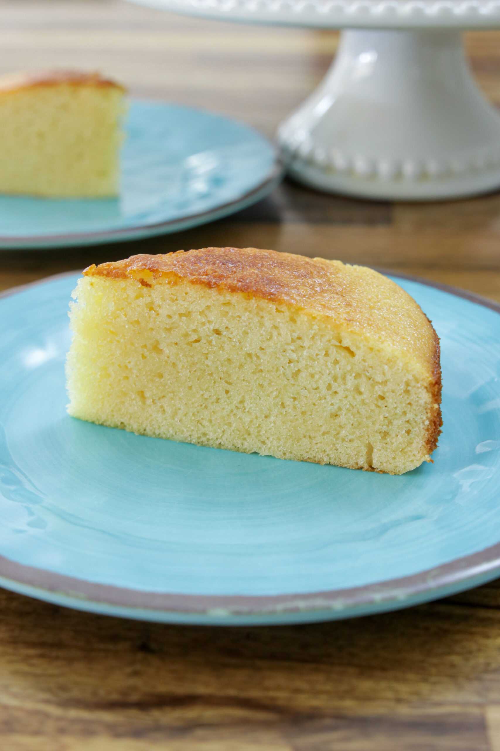 A single slice of condensed milk cake sits on a blue plate, with another plate and a cake stand blurred in the background.