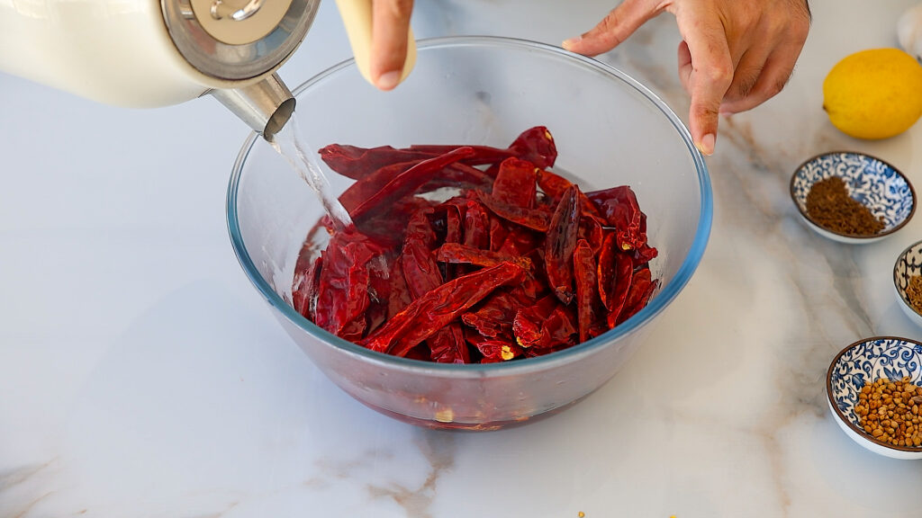 A person pours hot water from a kettle into a glass bowl filled with dried red chilies, surrounded by small bowls of spices and a lemon on a marble countertop.