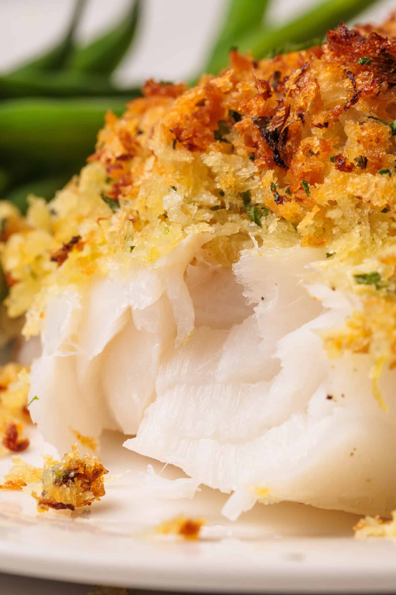 Close-up of a flaky baked panko cod fish fillet topped with a golden, crispy breadcrumb crust, with green vegetables blurred in the background.