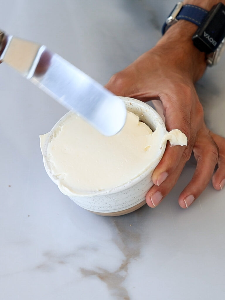 A person holding a small ceramic bowl filled with cream cheese while spreading it with an offset spatula.