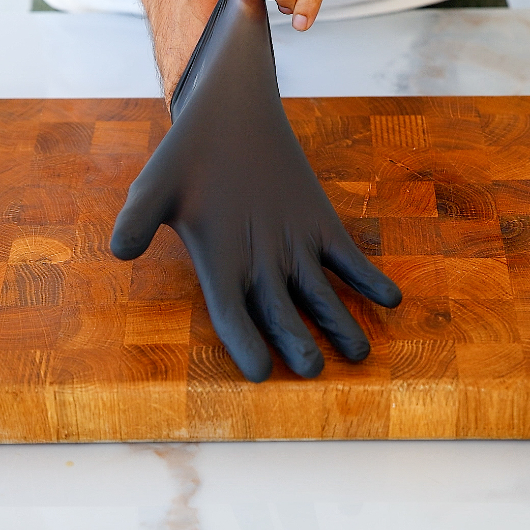 A person wearing a black disposable glove stands by a wooden cutting board, with their gloved hand resting flat on the board.