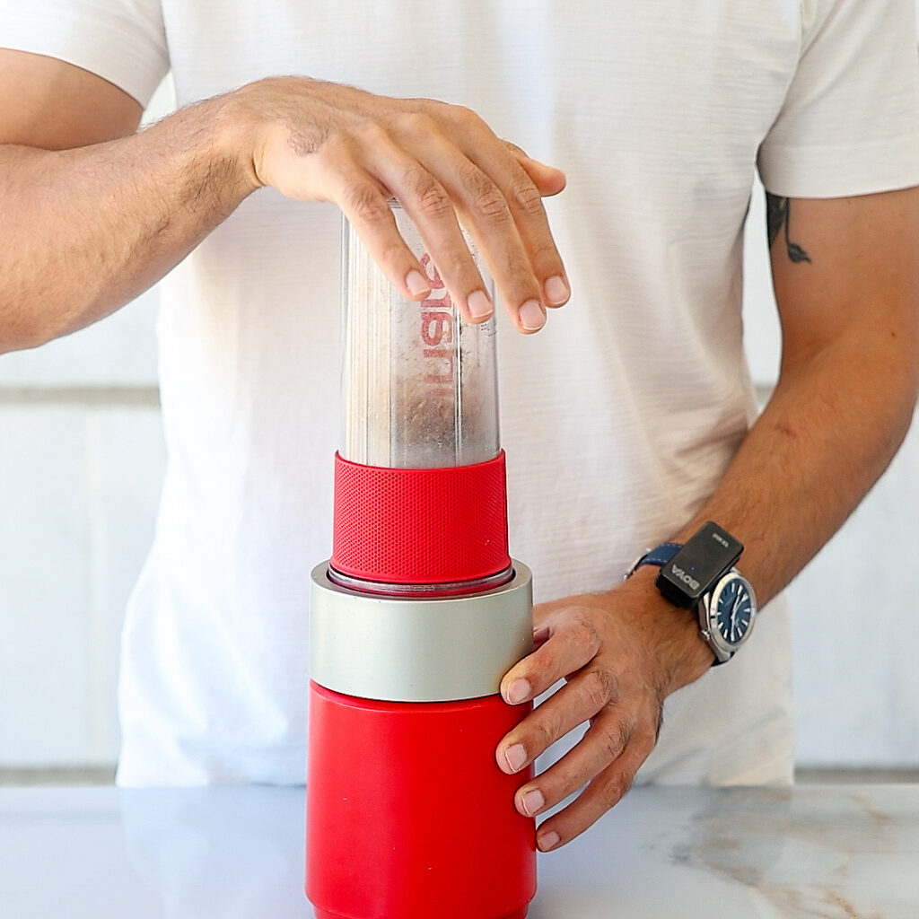 A person in a white shirt uses both hands to press down on a red and silver kitchen appliance, possibly a blender or juicer, placed on a counter. The person is wearing a wristwatch.
