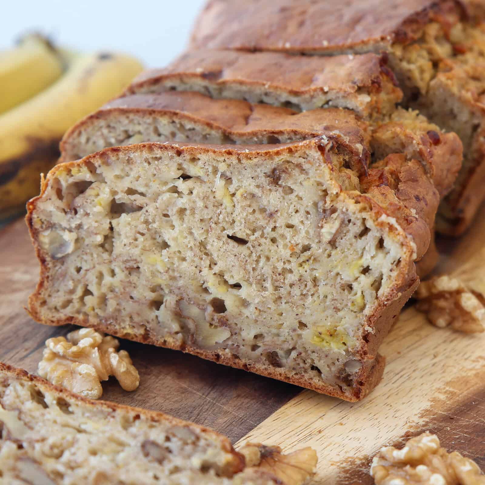 A close-up of sliced banana bread with cottage cheese and visible pieces of walnuts, resting on a wooden cutting board. Whole walnuts and bananas are in the background.