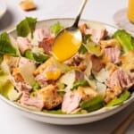 A hand drizzles yellow dressing from a spoon over a bowl of salad with romaine lettuce, croutons, chunks of tuna, and shaved parmesan cheese.