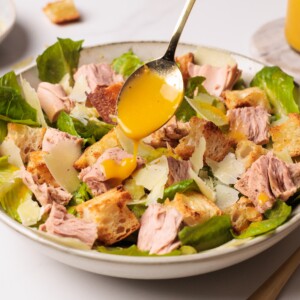 A hand drizzles yellow dressing from a spoon over a bowl of salad with romaine lettuce, croutons, chunks of tuna, and shaved parmesan cheese.