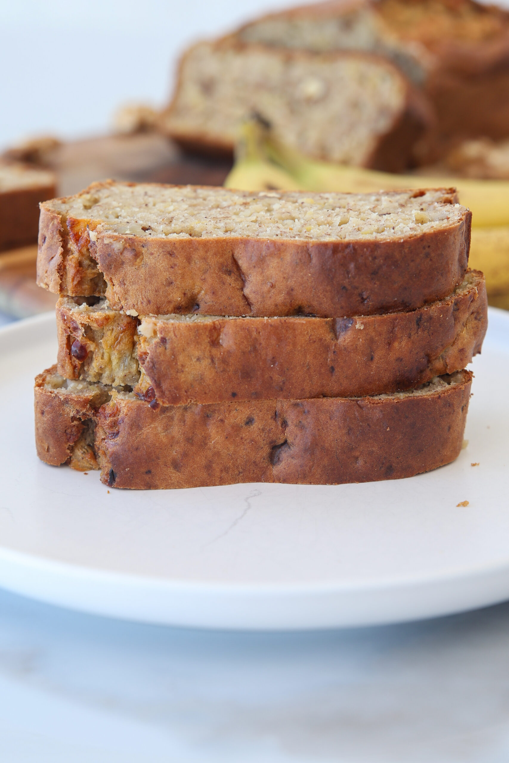 Three slices of healthy cottage cheese banana bread stacked on a white plate, with a loaf and bananas blurred in the background.