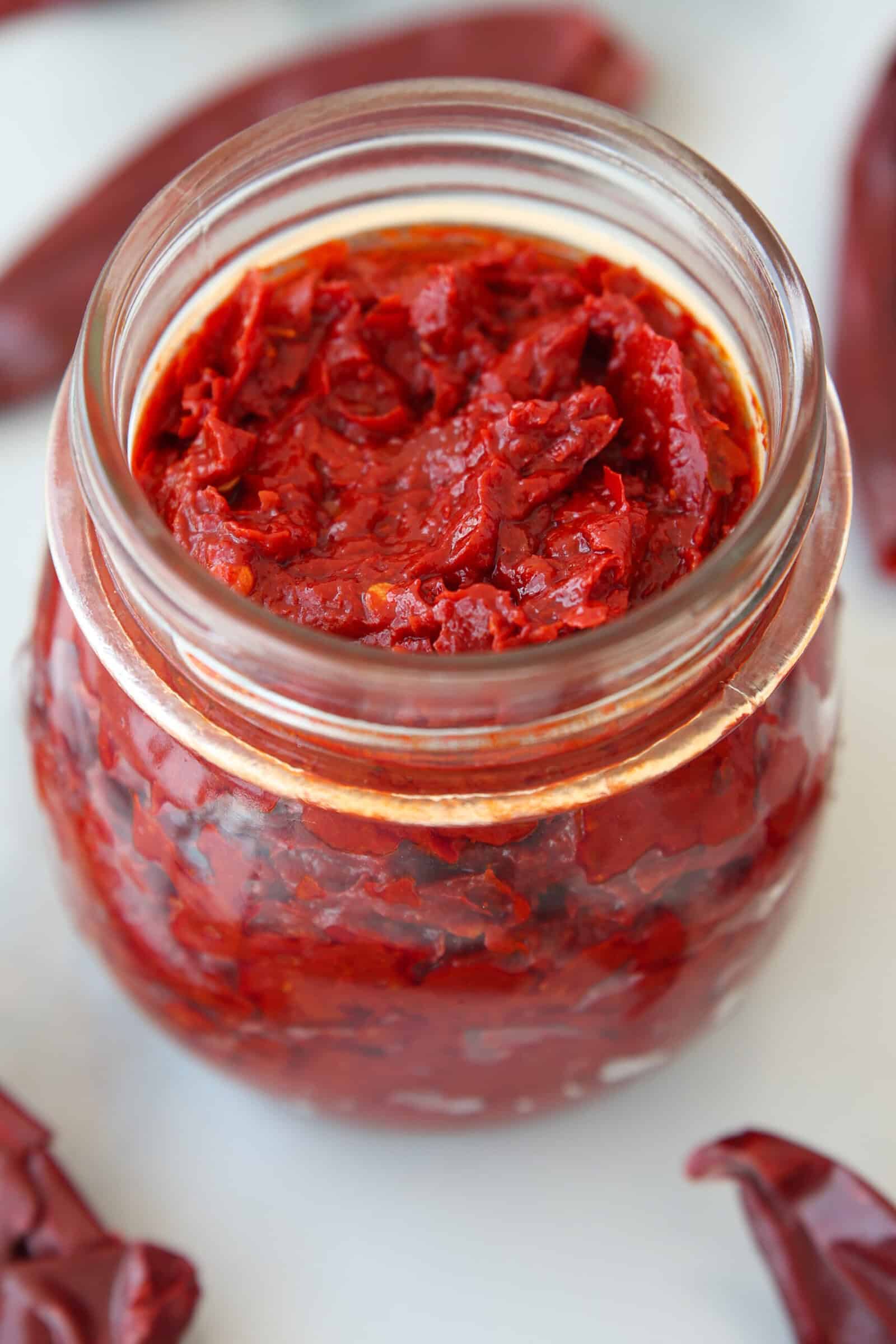 A glass jar filled with a chunky, bright red homemade harissa paste sits on a white surface, with dried red chili peppers scattered around it.