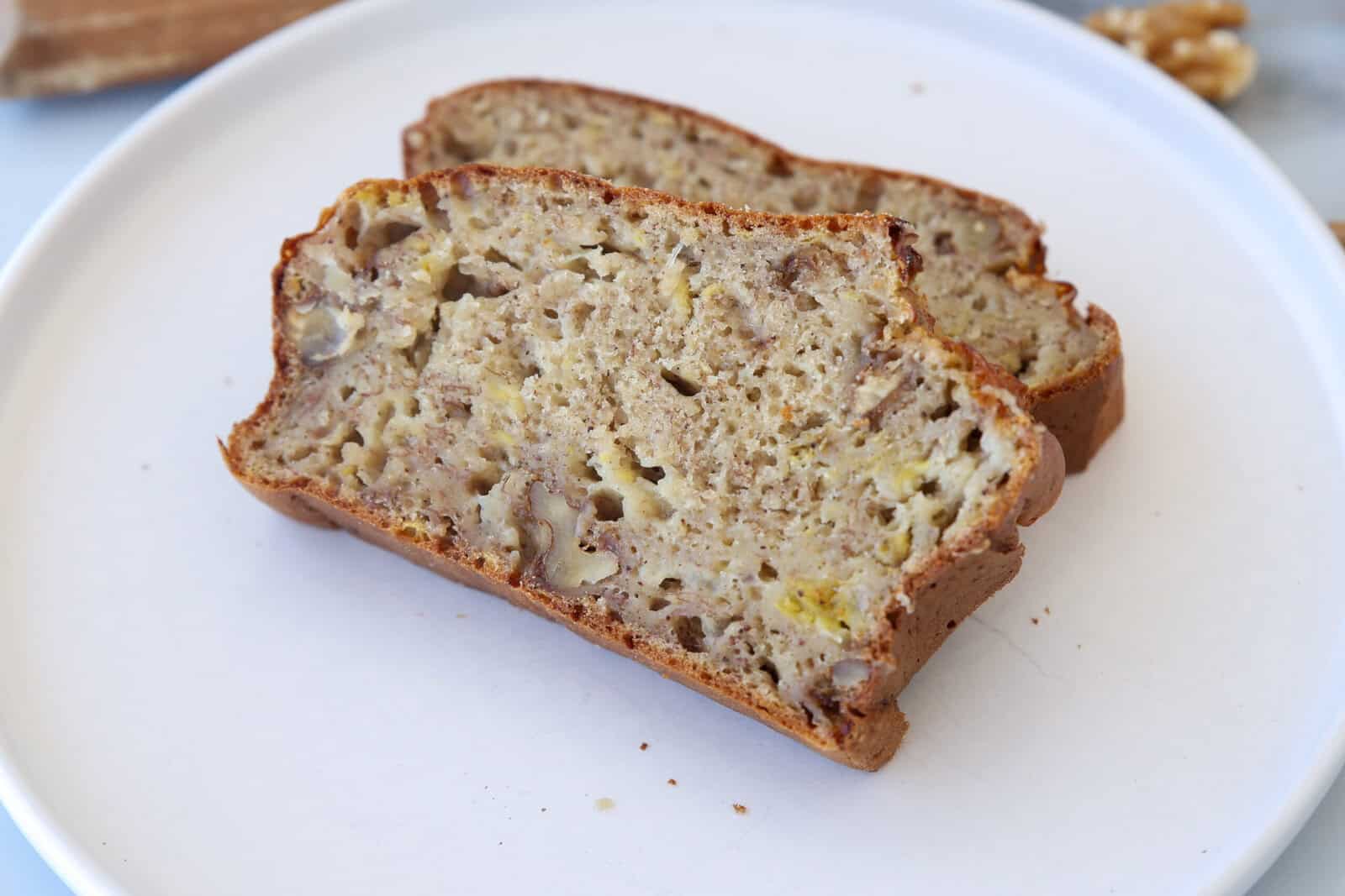 Two slices of high-protein banana nut bread with visible chunks of walnuts are placed on a white plate. The bread appears moist with a golden-brown crust.