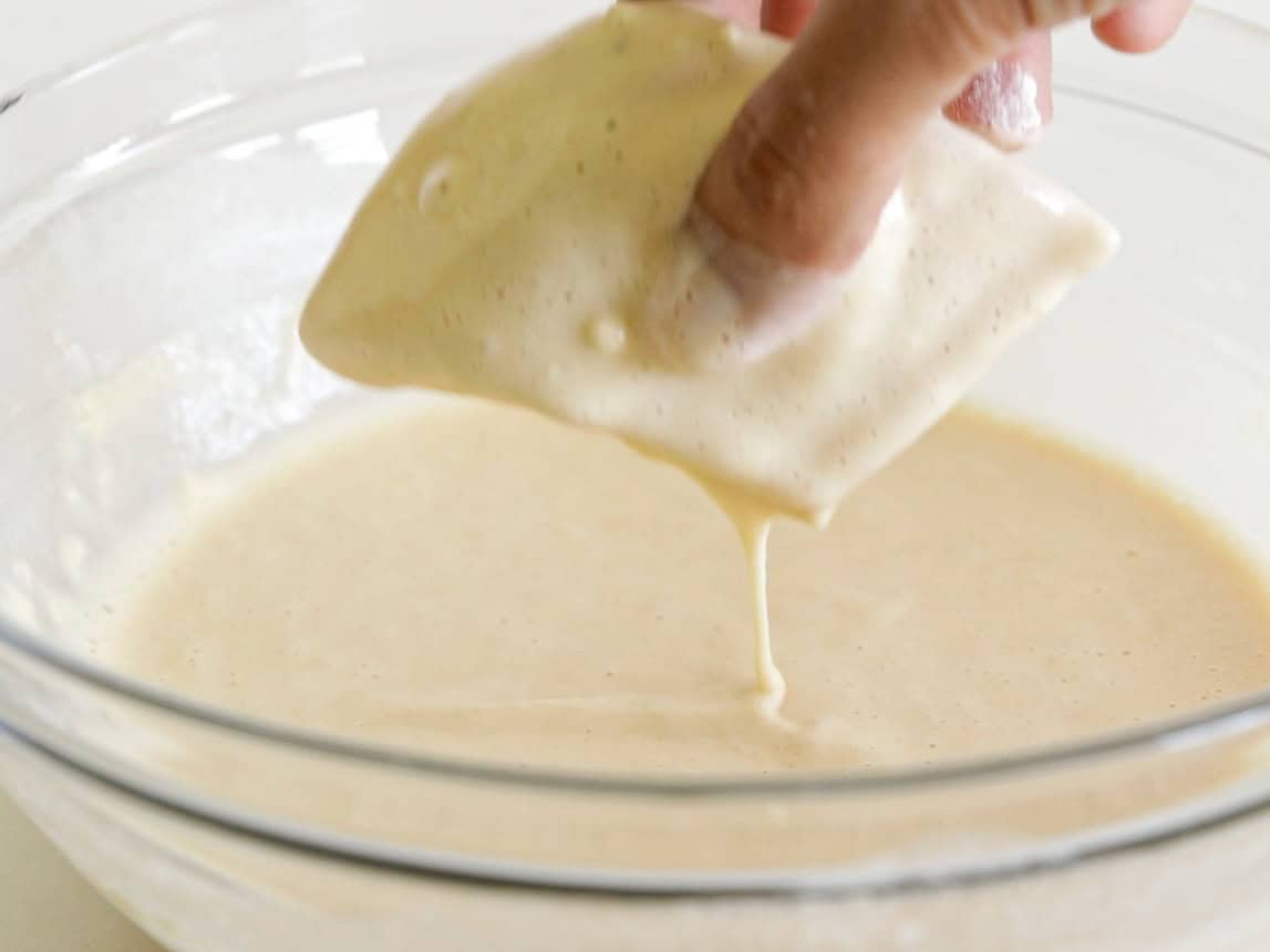 A hand dipping a piece of food into a thick, creamy batter in a clear glass bowl, with batter dripping back into the bowl.