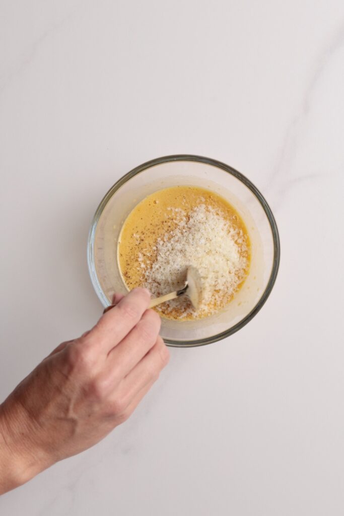 A hand stirring grated cheese into a bowl of beaten eggs with a spoon, on a white surface.