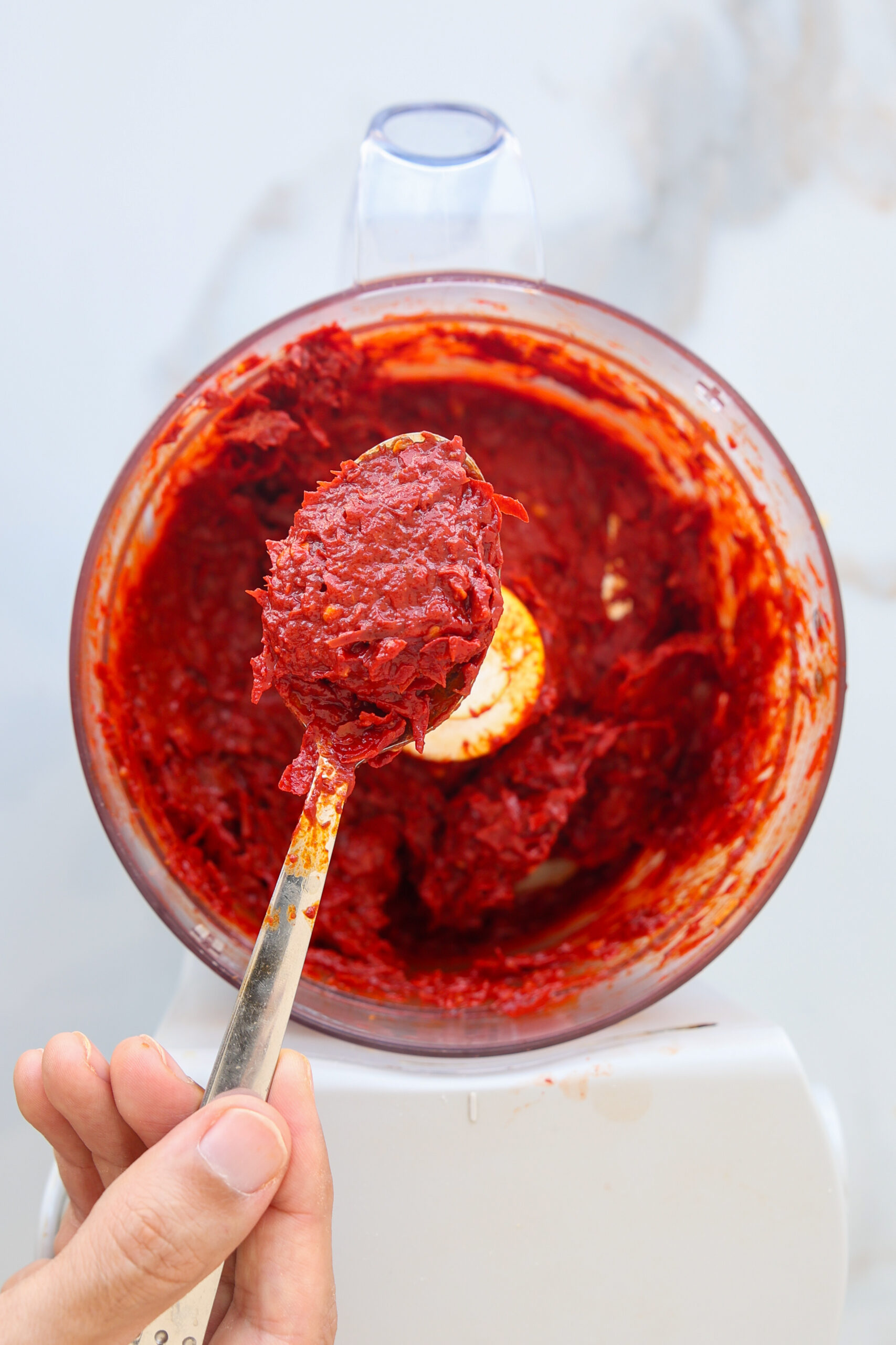 A hand holding a spoonful of thick red chili paste over a food processor bowl filled with the same mixture, likely a homemade sauce or condiment.
