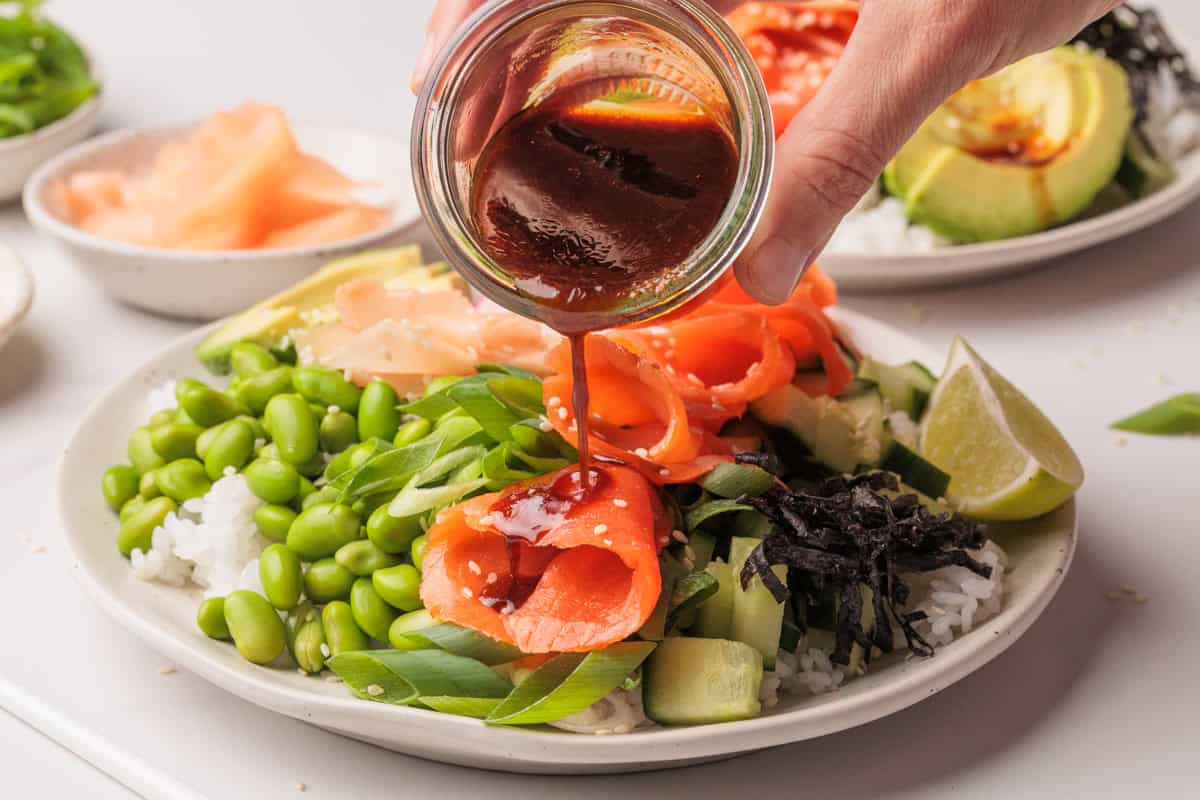 A hand pours dark sauce from a jar onto a smoked salmon poke bowl filled with rice, sliced salmon, edamame, seaweed, cucumber, scallions, pickled ginger, and lime wedges on a white plate.