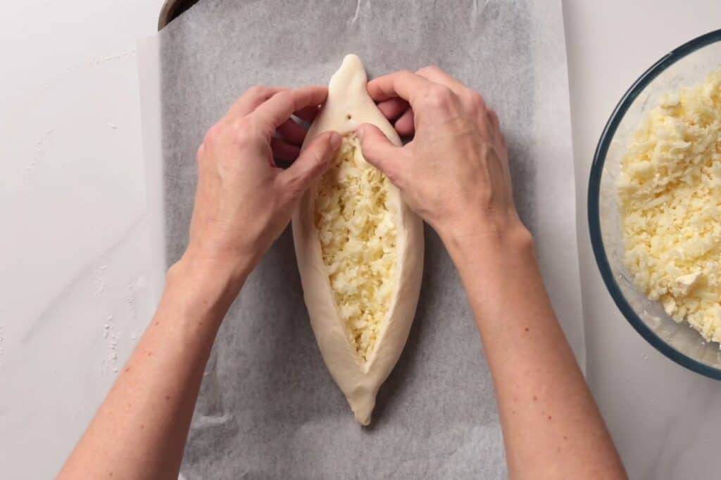 Two hands shape a boat-like dough filled with shredded cheese on parchment paper, with a glass bowl of more cheese nearby on a white countertop.