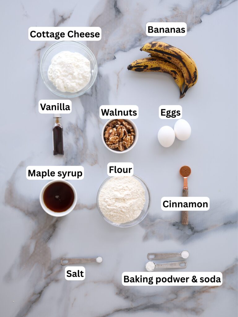 Ingredients for baking are arranged on a marble surface, including cottage cheese, ripe bananas, vanilla, walnuts, eggs, maple syrup, flour, cinnamon, salt, and baking powder & soda, each labeled with text.