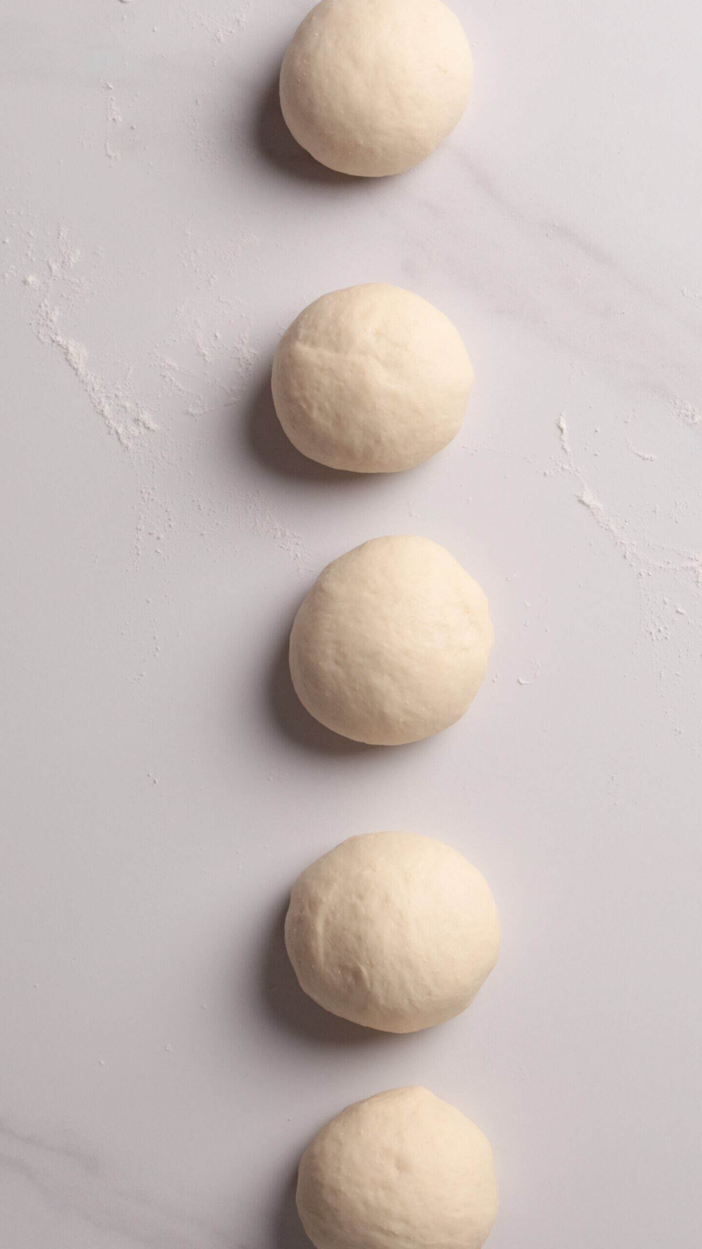 Five round balls of dough are evenly spaced in a vertical line on a light-colored, lightly floured surface.