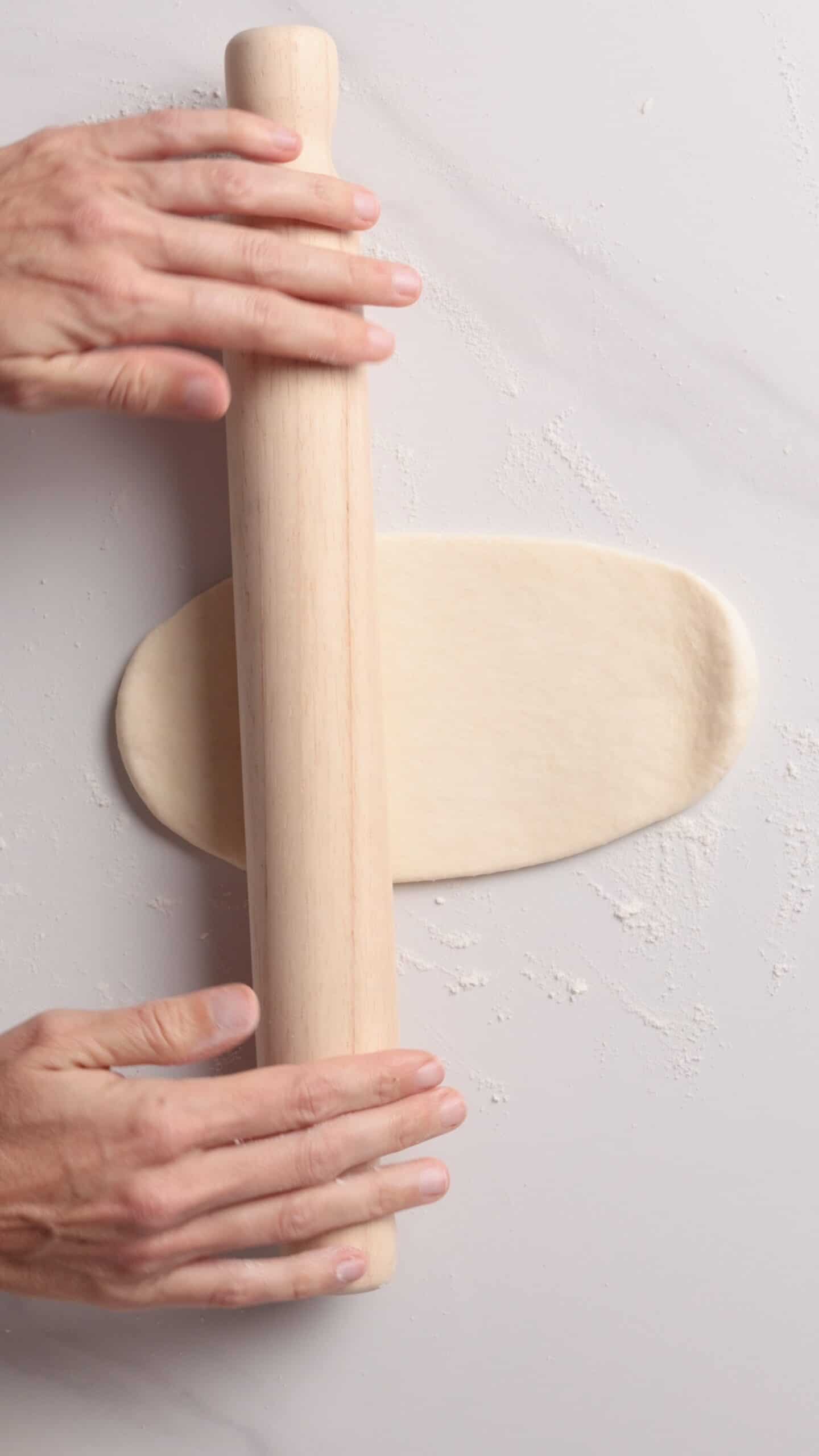 Two hands use a wooden rolling pin to flatten a piece of dough on a floured white surface.
