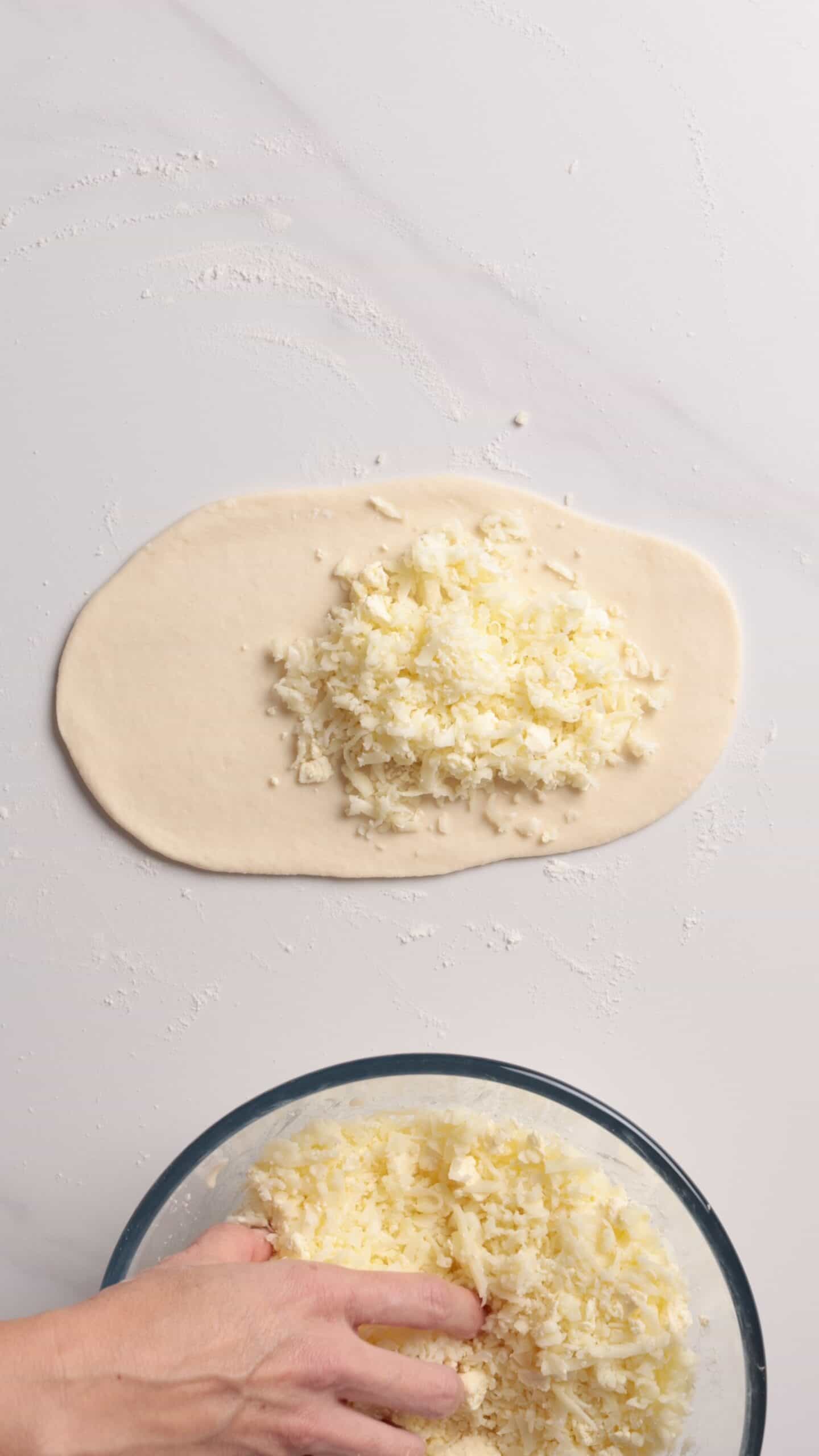 A hand reaches into a bowl of shredded cheese, while a portion of dough on a floured surface is topped with a pile of cheese.