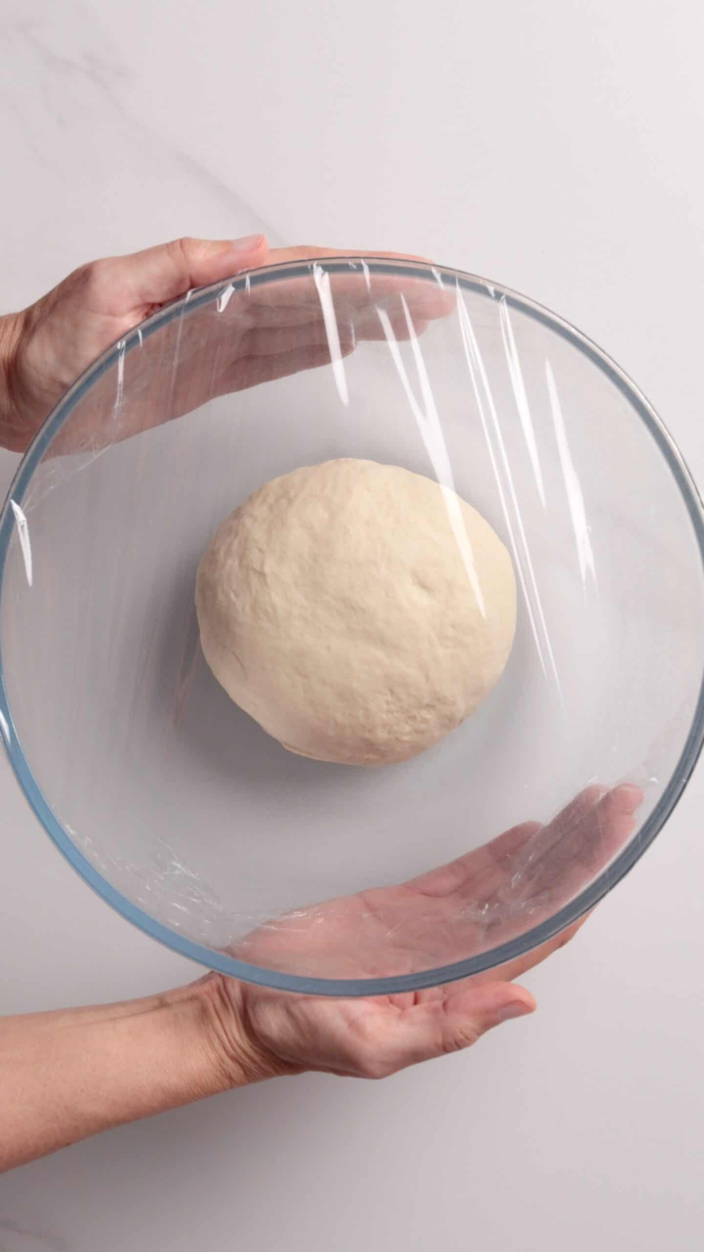 A person's hands holding a glass bowl with dough inside, covered with plastic wrap, ready for proofing on a light surface.