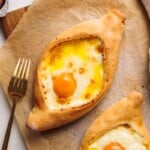 Two khachapuri breads on parchment paper, each shaped like a boat and filled with melted cheese and a baked egg yolk in the center. A gold fork is placed to the left side of the bread.