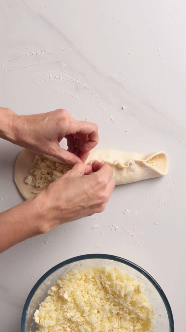 Hands fold dough around cheese filling on a floured surface; a bowl with grated cheese sits nearby.