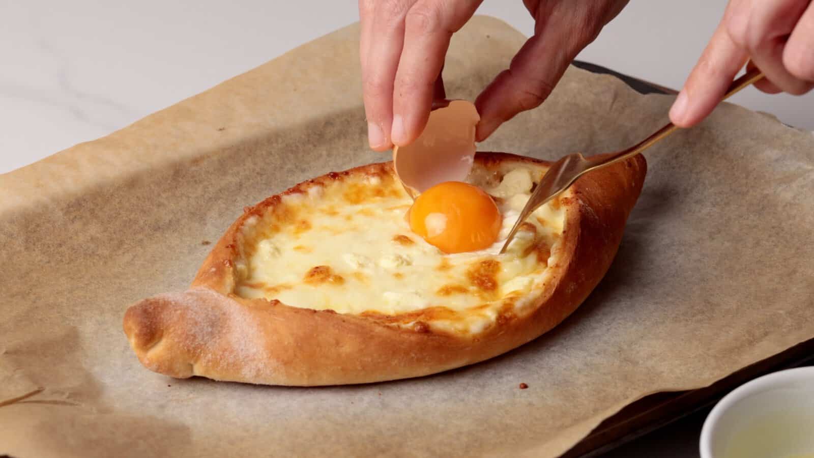 A hand cracks a raw egg onto a baked khachapuri, a cheese-filled bread, on parchment paper, with a small bowl nearby.