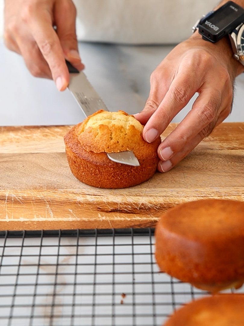 A person slices a small round cake on a wooden cutting board. Another cake is nearby on a cooling rack. The person wears a watch and uses a serrated knife to cut.