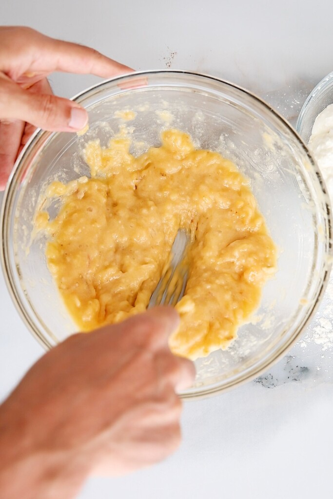 A person mashing bananas in a glass bowl with a fork. Another glass bowl with white flour is partially visible on the side. The scene is set on a light-colored surface.