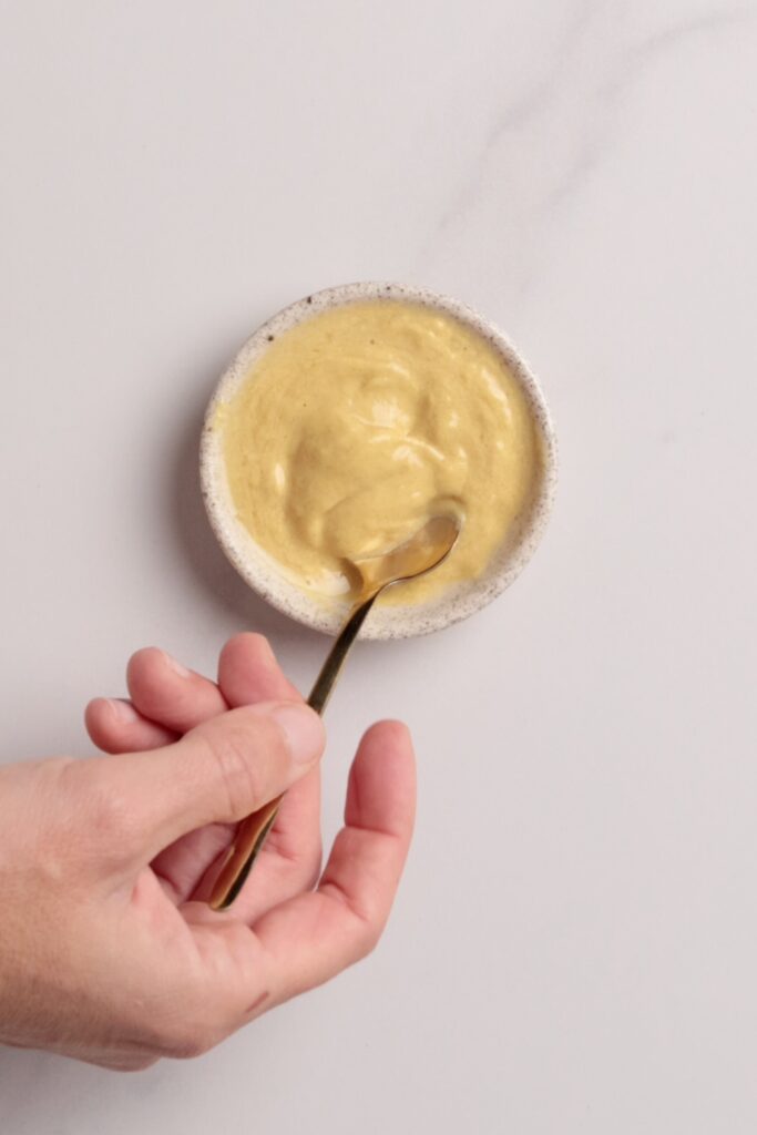A hand holding a small bowl of Dijon mustard and lemon juice, with a spoon resting inside, on a light-colored surface.