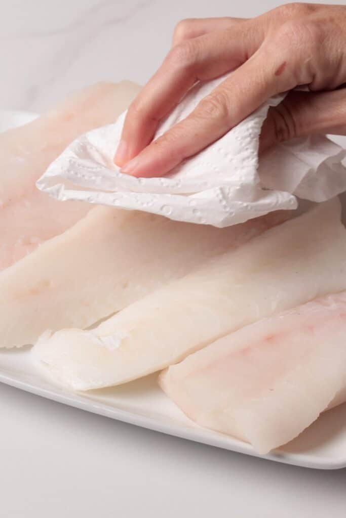 A hand uses a paper towel to pat dry raw cod fish fillets arranged on a white plate, on a light-colored surface.