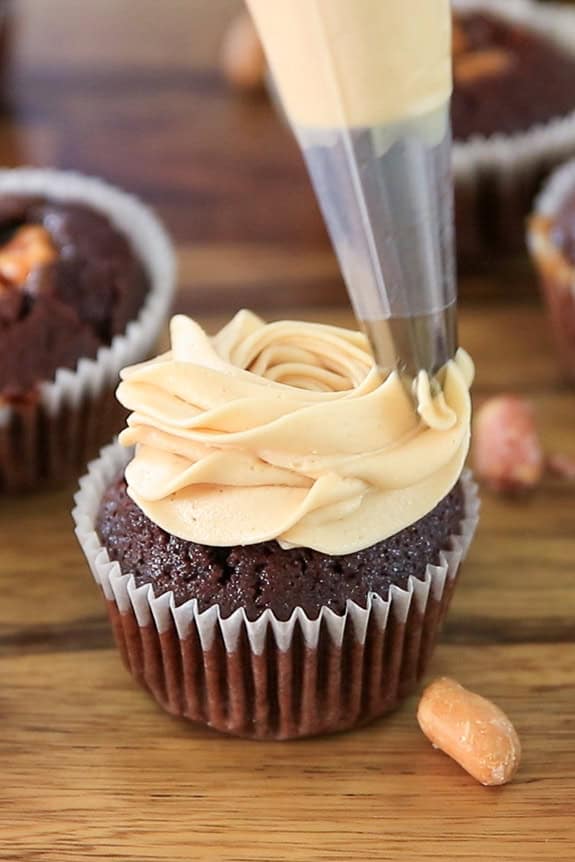 A chocolate cupcake in a white paper liner is being topped with swirled light brown frosting from a piping bag. Peanuts and other cupcakes are visible in the background on a wooden surface.
