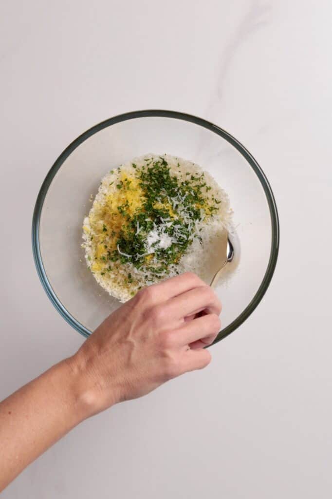 A hand holding a spoon is mixing grated parmesan, chopped herbs, and panko breadcrumbs in a clear glass bowl on a white surface.