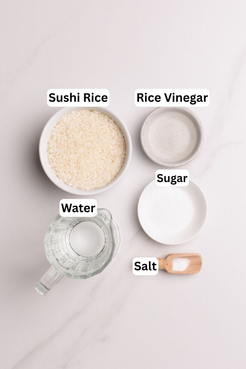 A bowl of sushi rice, a small bowl of rice vinegar, a plate of sugar, a wooden spoon with salt, and a glass jug of water are arranged on a white surface, each labeled with its name.