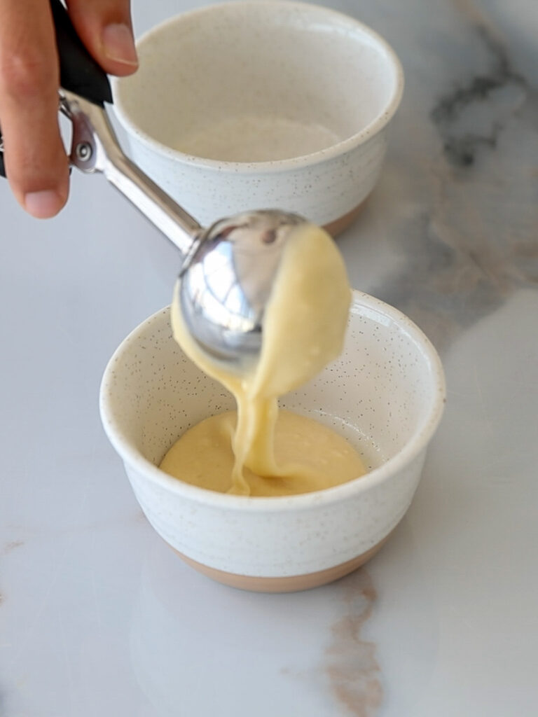 A hand uses an ice cream scoop to pour thick cake batter into a small white ceramic bowl on a marble surface, with another empty bowl in the background.